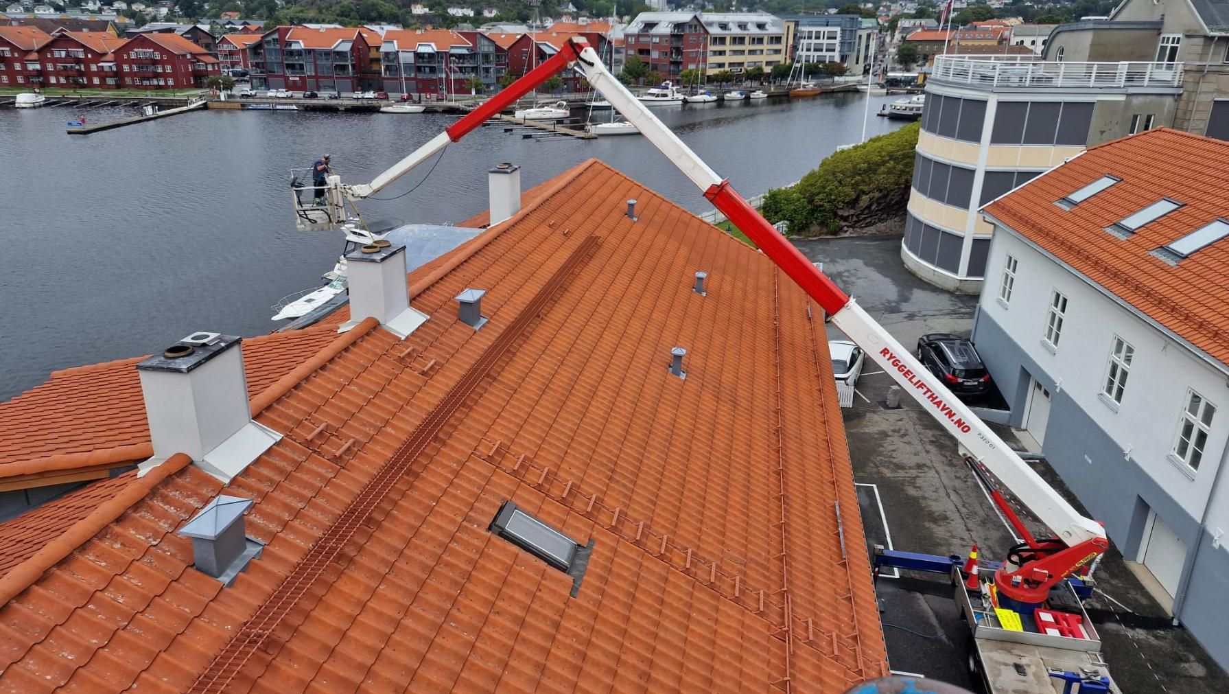 Liftbil med lang arm utfører arbeid over tak på bygg ved havneområde.