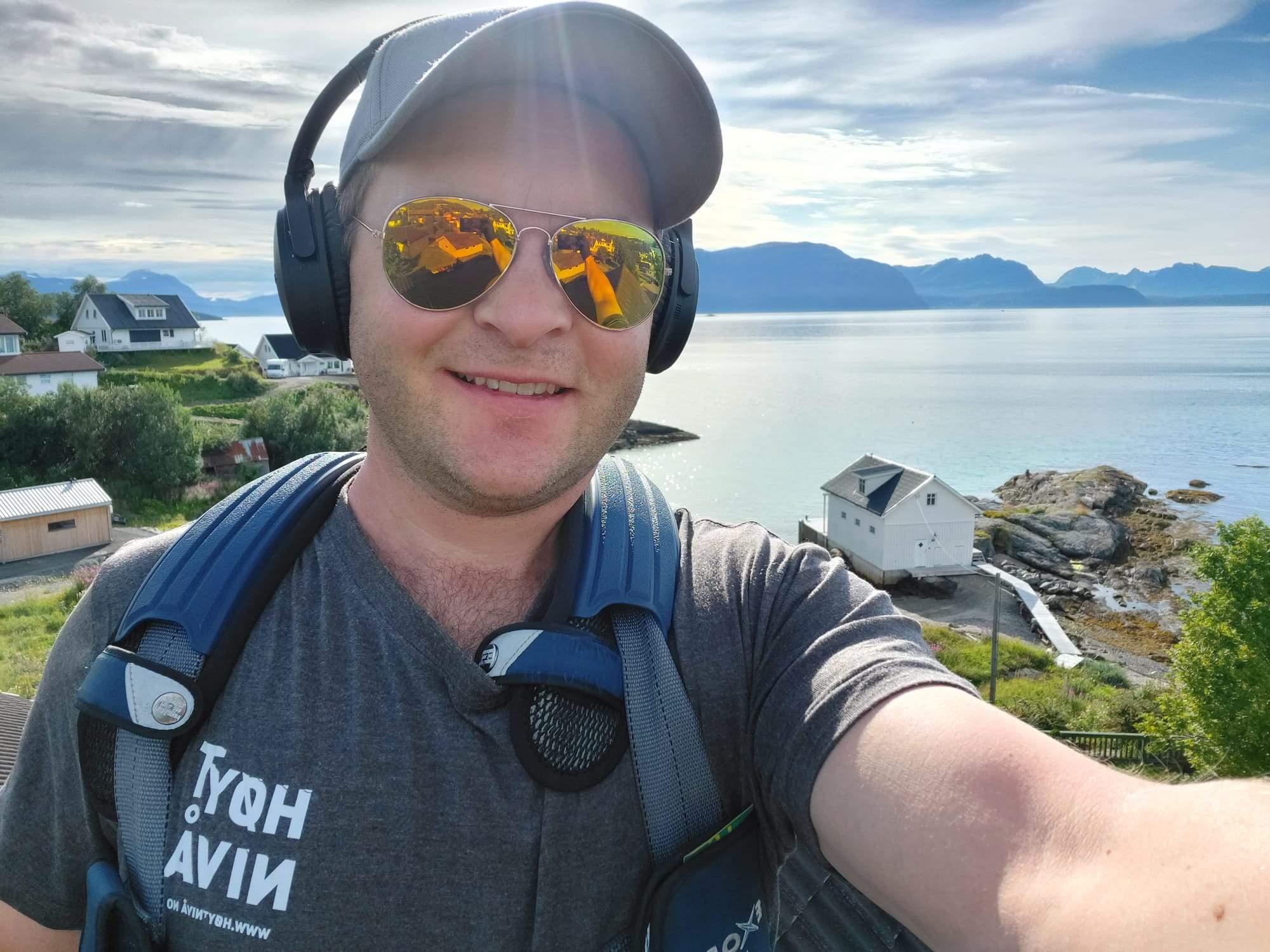 Ansatt i Høyt Nivå tar selfie utendørs med sjøutsikt og fjell i bakgrunnen. Symboliserer arbeidsmiljø og ulike arbeidslokasjoner.